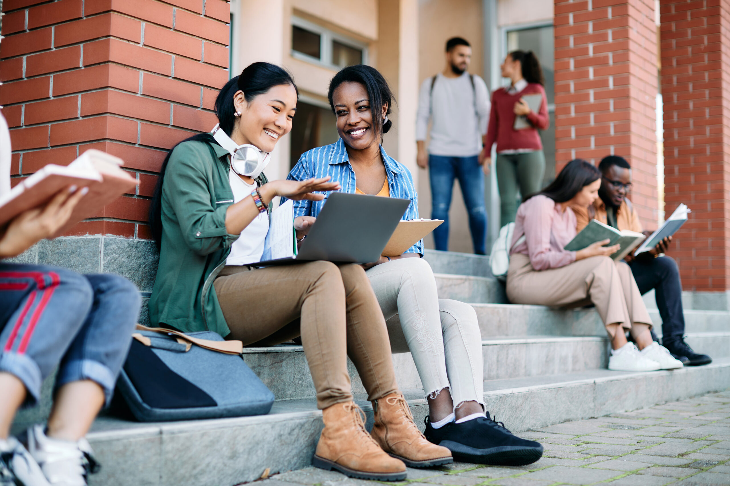 Students smiling while using laptop in front of school building.
