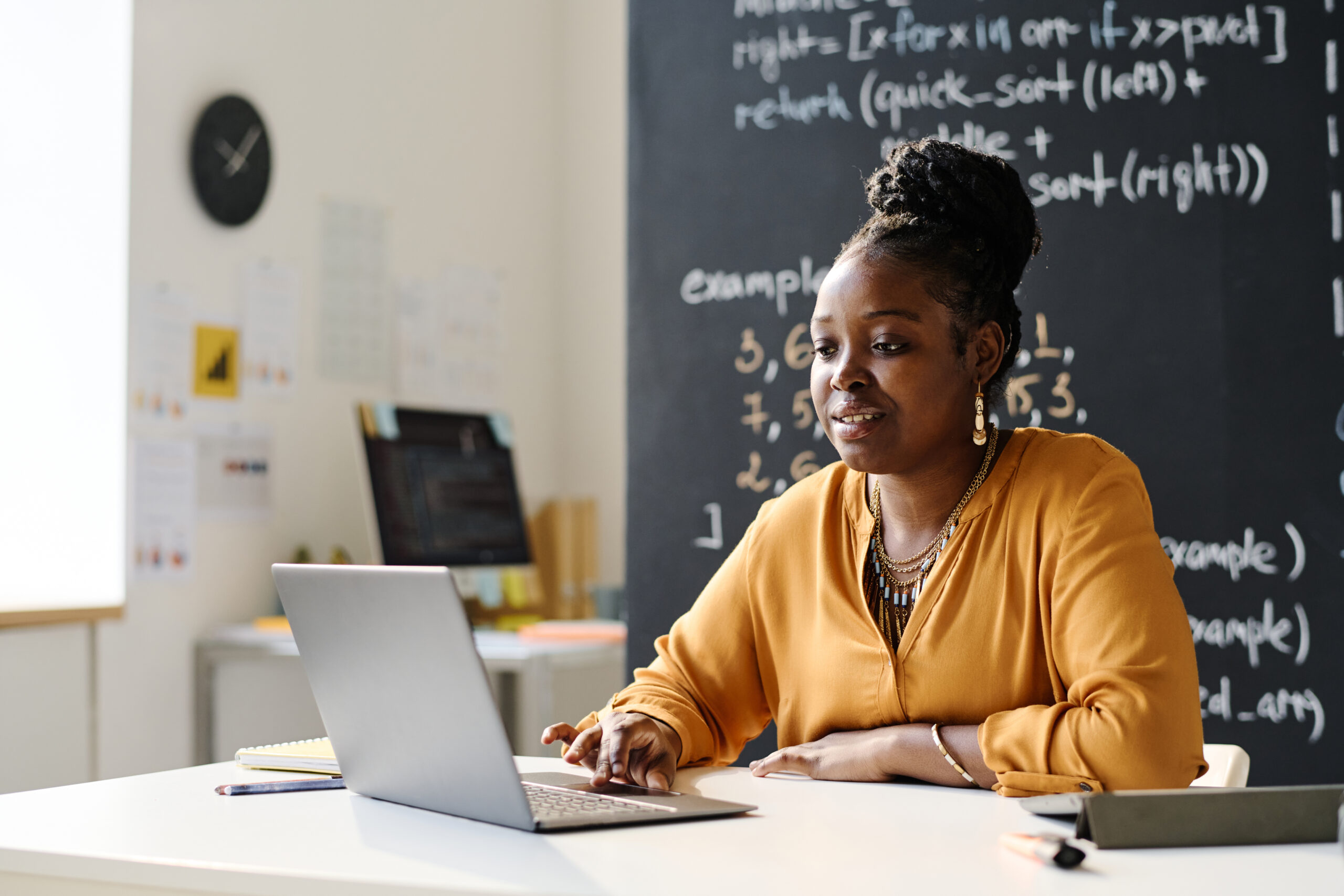 Teacher working on laptop at desk in front of chalkboard.